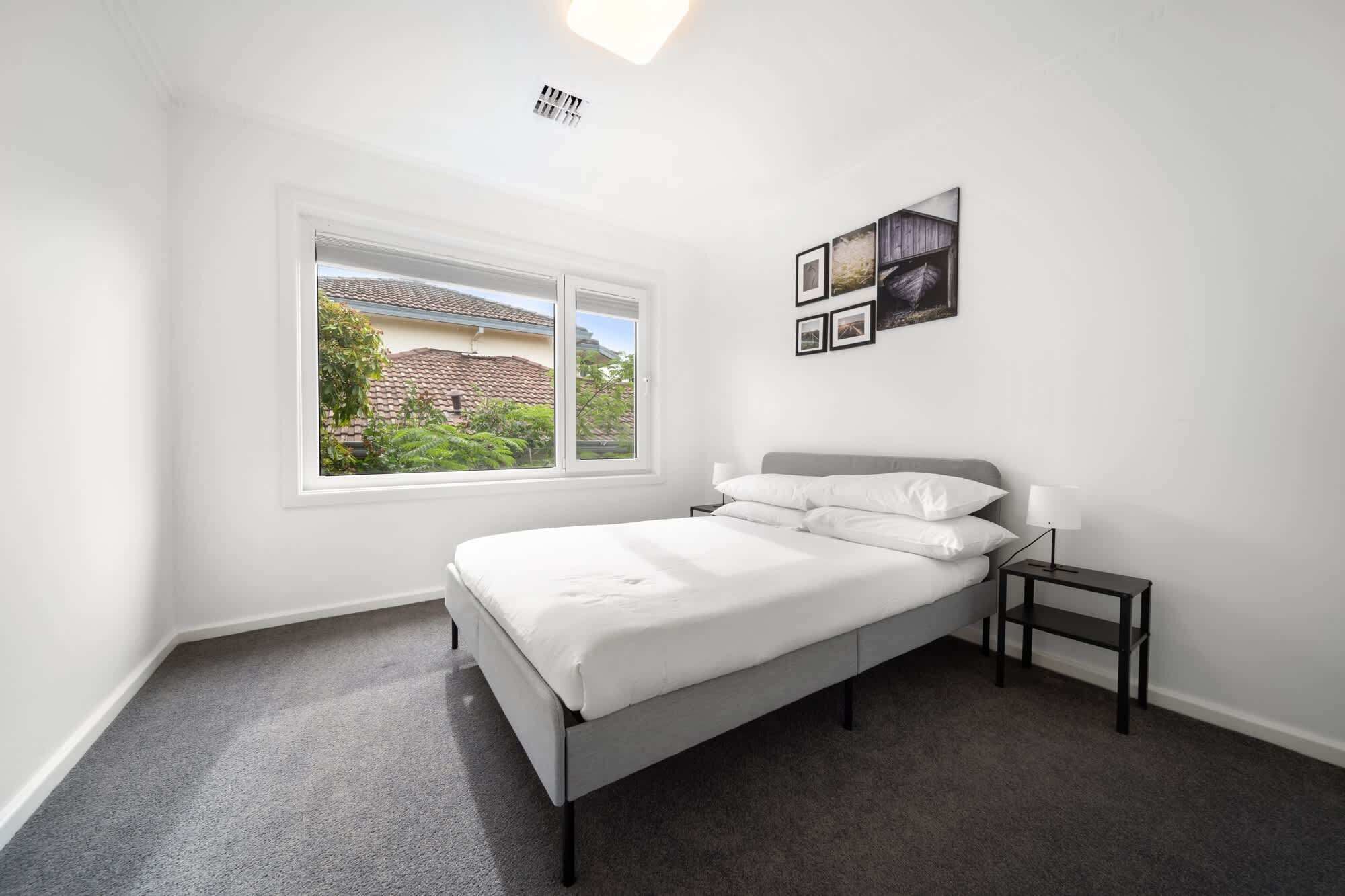 Une chambre à coucher avec un lit, une table de chevet, une lampe et une fenêtre donnant sur une maison et de la verdure.