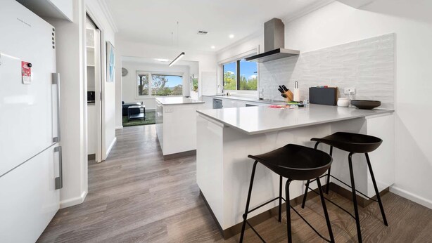 A modern kitchen with white cabinetry, a central island, and two bar stools. - 4-Bed Family Home with Spacious Backyard and BBQ (Curtin)