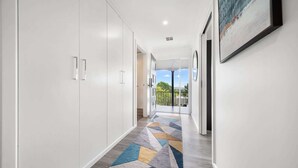 A modern hallway with white walls, a geometric patterned rug, and a view of a balcony with greenery. - 4-Bed Family Home with Spacious Backyard and BBQ (Curtin)