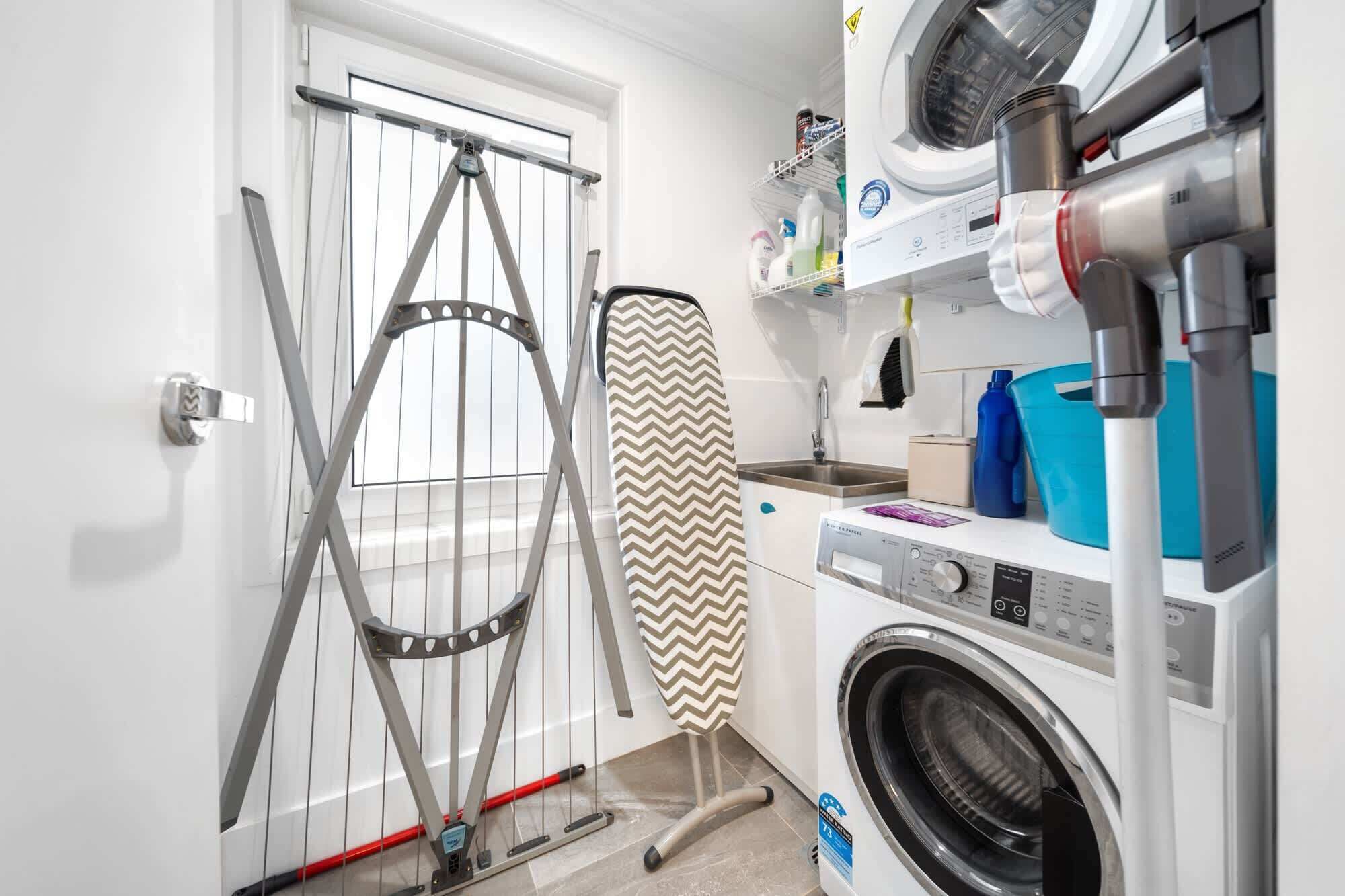 Een wasruimte met een wasmachine, droger, strijkplank en een droogrek.