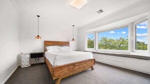 A bedroom with a wooden bed, white walls, a window with a view of trees, and a small desk with a lamp. - 4-Bed Family Home with Spacious Backyard and BBQ (Curtin)