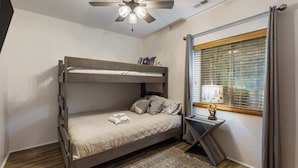 Un lit superposé avec un ventilateur de plafond, une fenêtre avec des stores et une petite table de chevet avec une lampe.