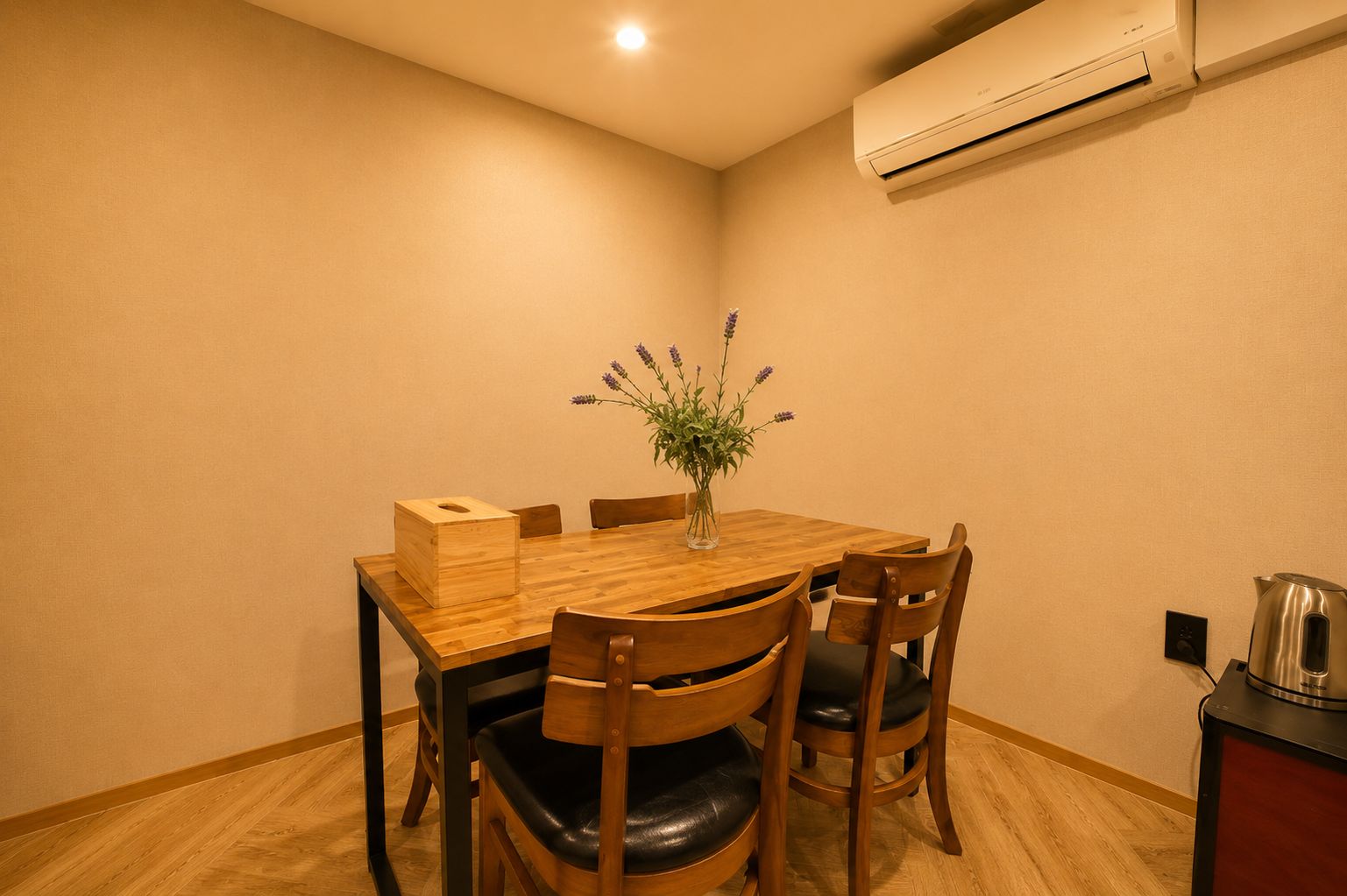 Un pequeño comedor con una mesa de madera, cuatro sillas, un florero con flores y un equipo de aire acondicionado en la pared.