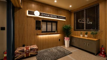 A hotel reception area with a wooden panel wall, a reception desk, a potted plant, a fire extinguisher, and a fire hose.
