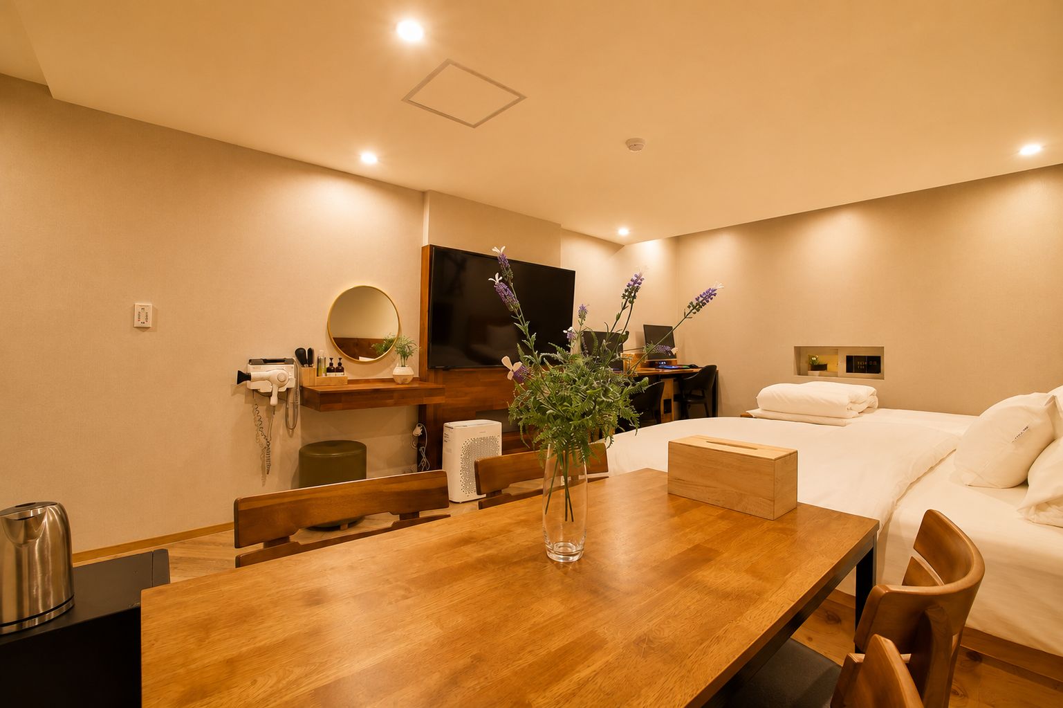 A hotel room with a wooden dining table, a bed with white linens, a flat-screen TV mounted on the wall, and a small desk with a computer.