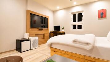 A modern hotel room with a large bed, a flat-screen TV, a desk with a computer, and a small kitchenette area.