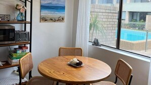 Uma área de refeições com uma mesa de madeira redonda, quatro cadeiras de vime, uma estante com livros e objetos decorativos, um quadro com uma paisagem de praia e uma janela com vista para uma piscina.
