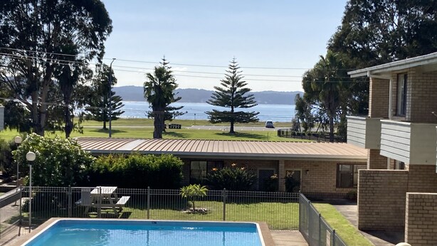 Piscina com vista para o mar e montanhas, rodeada por um jardim cercado com um edifício e árvores.