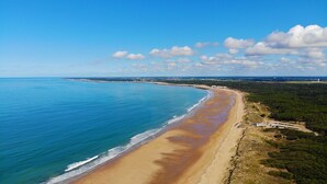 Une vue aérienne d’une plage avec une longue étendue de sable, une mer bleue et limpide, et une zone boisée en arrière-plan.