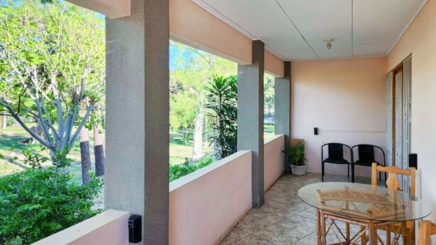 Un espace extérieur abrité, doté d’une table et de chaises en verre, entouré de murs et de colonnes, et offrant une vue sur un jardin avec des arbres et des plantes.