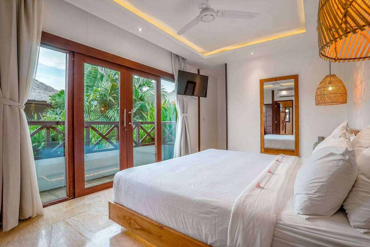 Un dormitorio con una cama grande, ventilador de techo, televisor y balcón con vistas a zonas verdes.