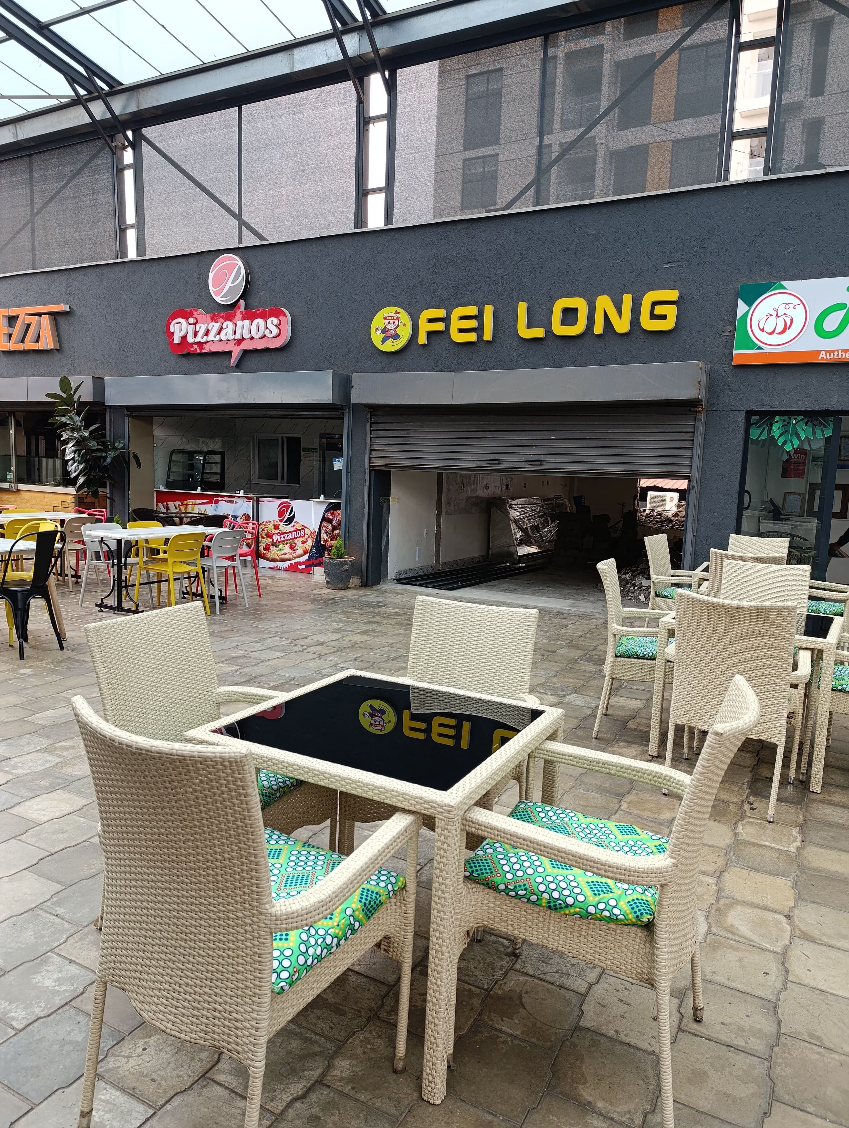 An outdoor seating area with wicker chairs and a table, in front of a building with a 'Pizzanos' sign and a 'FEI LONG' sign.