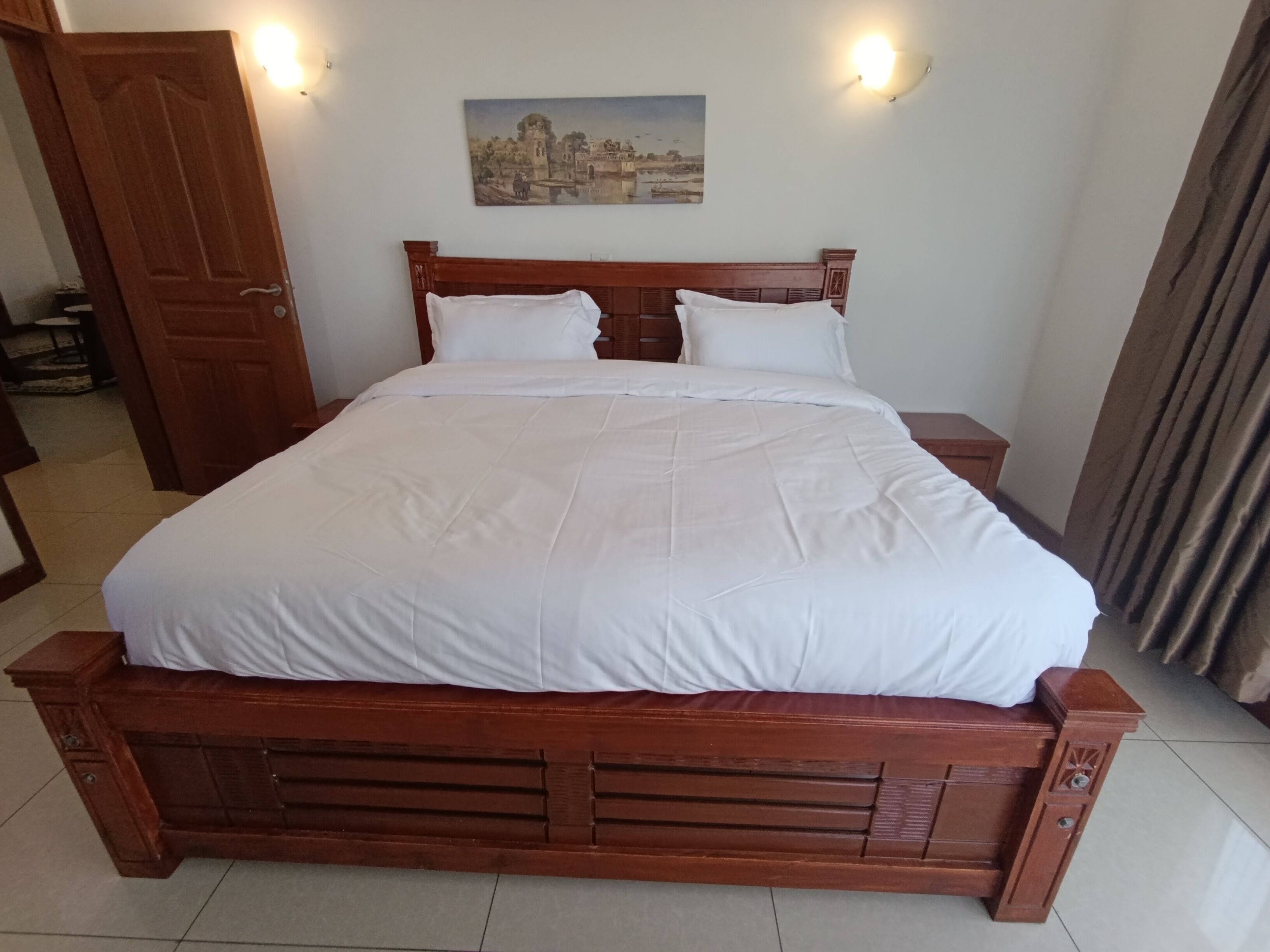 Un lit en bois avec une literie blanche, une table de chevet, une lampe fixée au mur et un tableau au-dessus du lit.