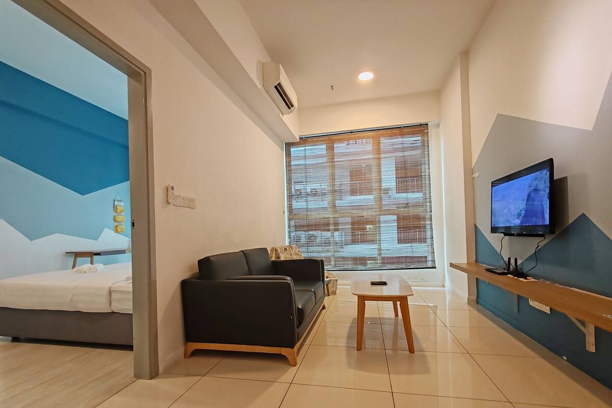 Habitació d'hotel moderna amb llit, sofà, taula petita, televisió i finestra gran amb persianes.