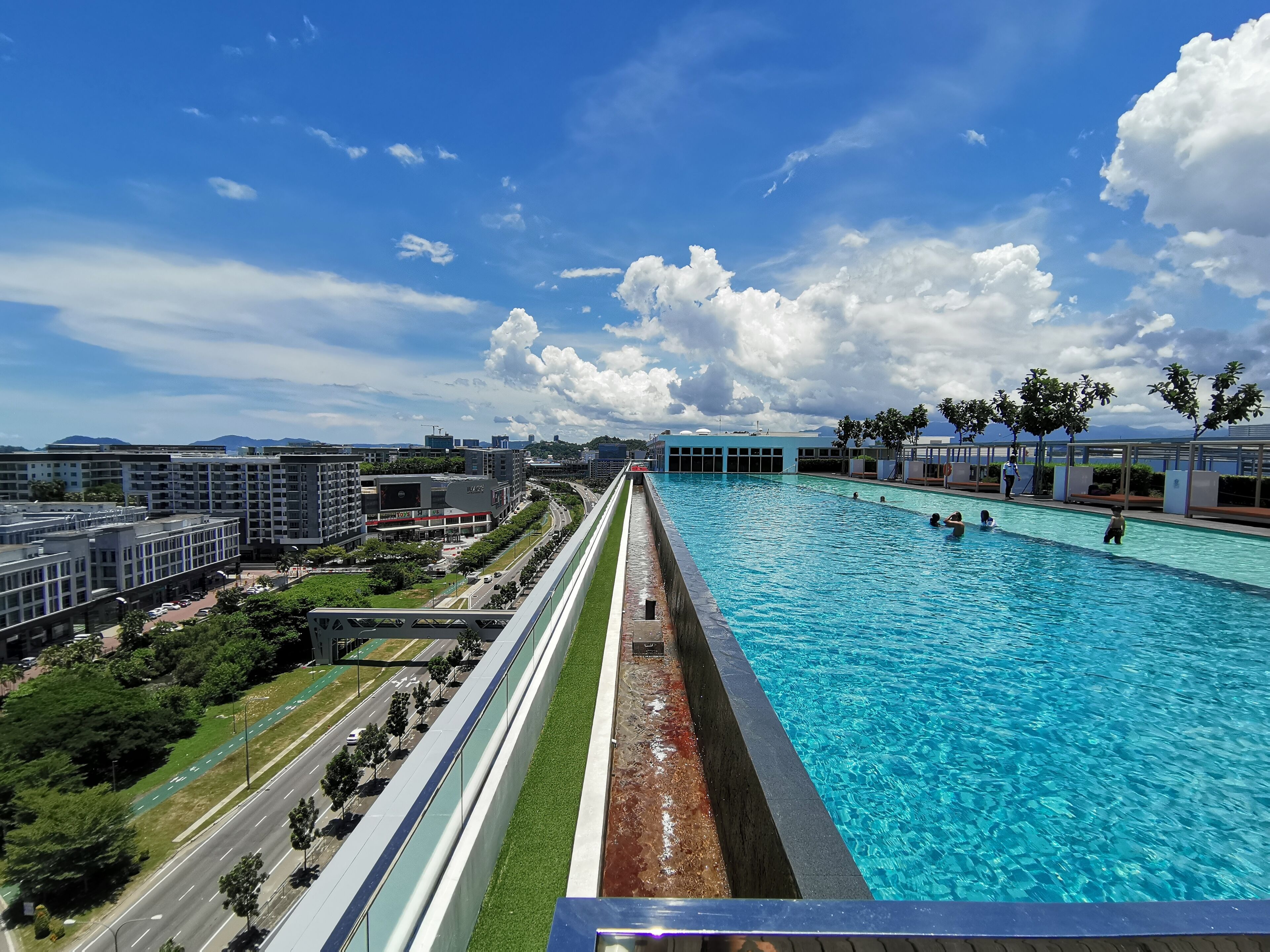 A large swimming pool with clear blue water, surrounded by modern buildings and a road.