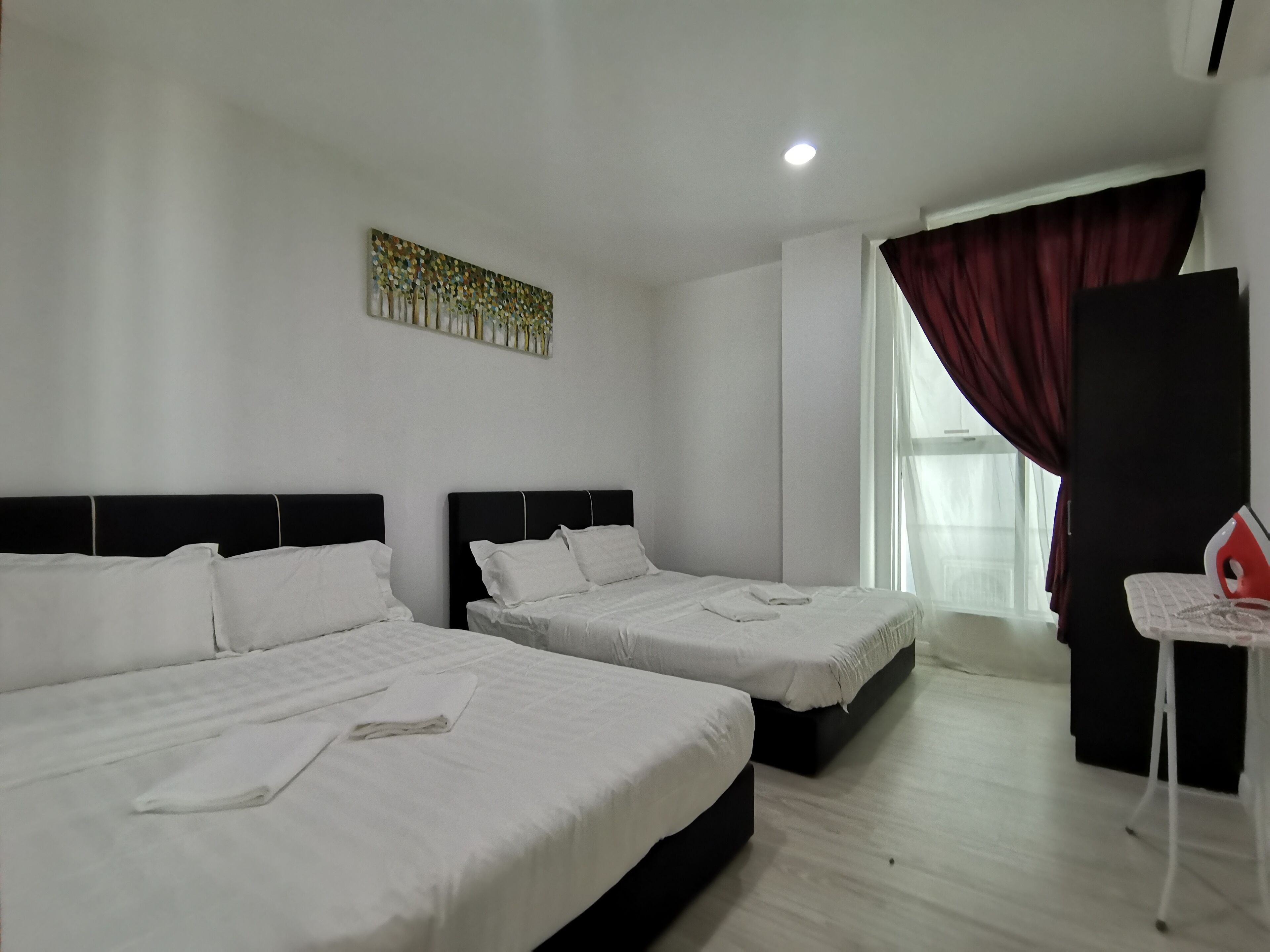 A hotel room with two beds, a ceiling light, a window with curtains, and a small table with an iron.