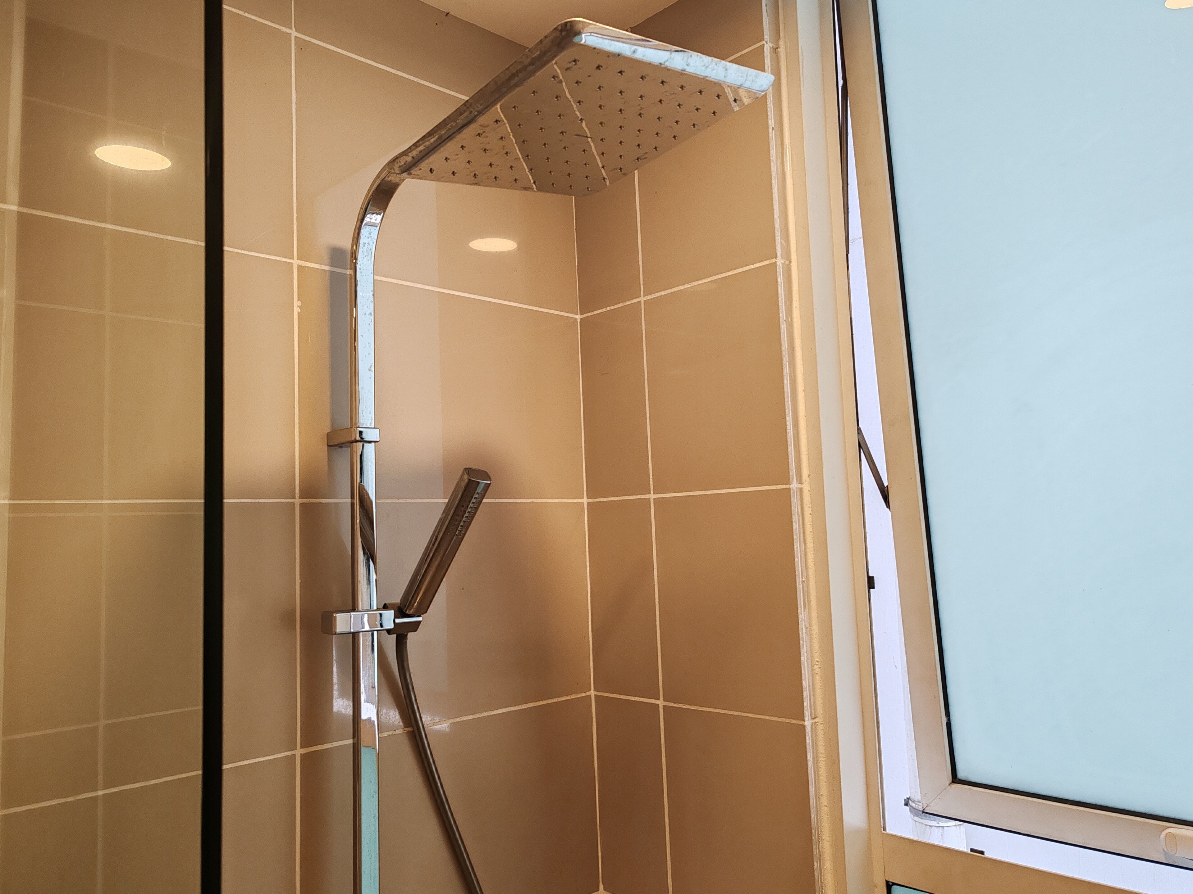 A shower with a handheld showerhead and a wall-mounted rain showerhead in a room with beige tiled walls and a window.