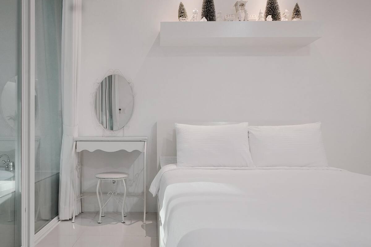 Une chambre à coucher moderne, aux tons blancs, avec une douche en verre, un miroir rond, une petite table de chevet et un lit avec une literie blanche.