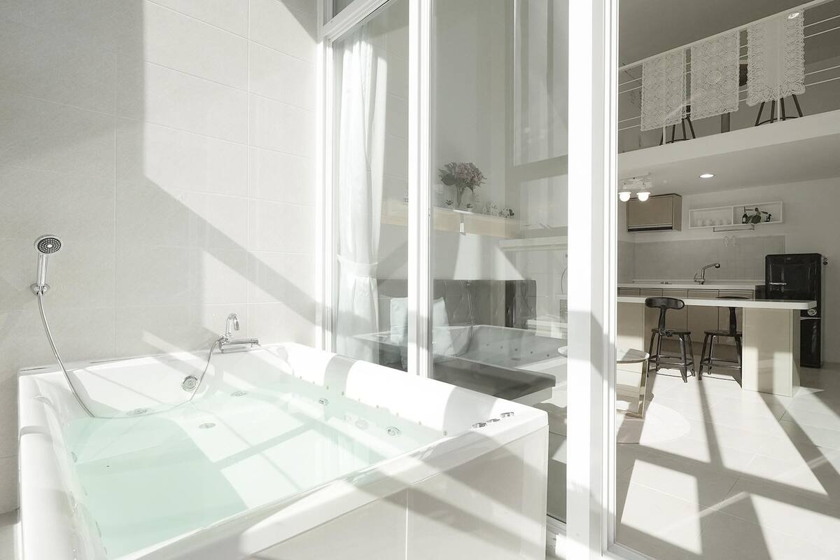 Une salle de bain moderne avec une baignoire entourée de parois en verre, une douche et un coin cuisine visible à travers le verre.