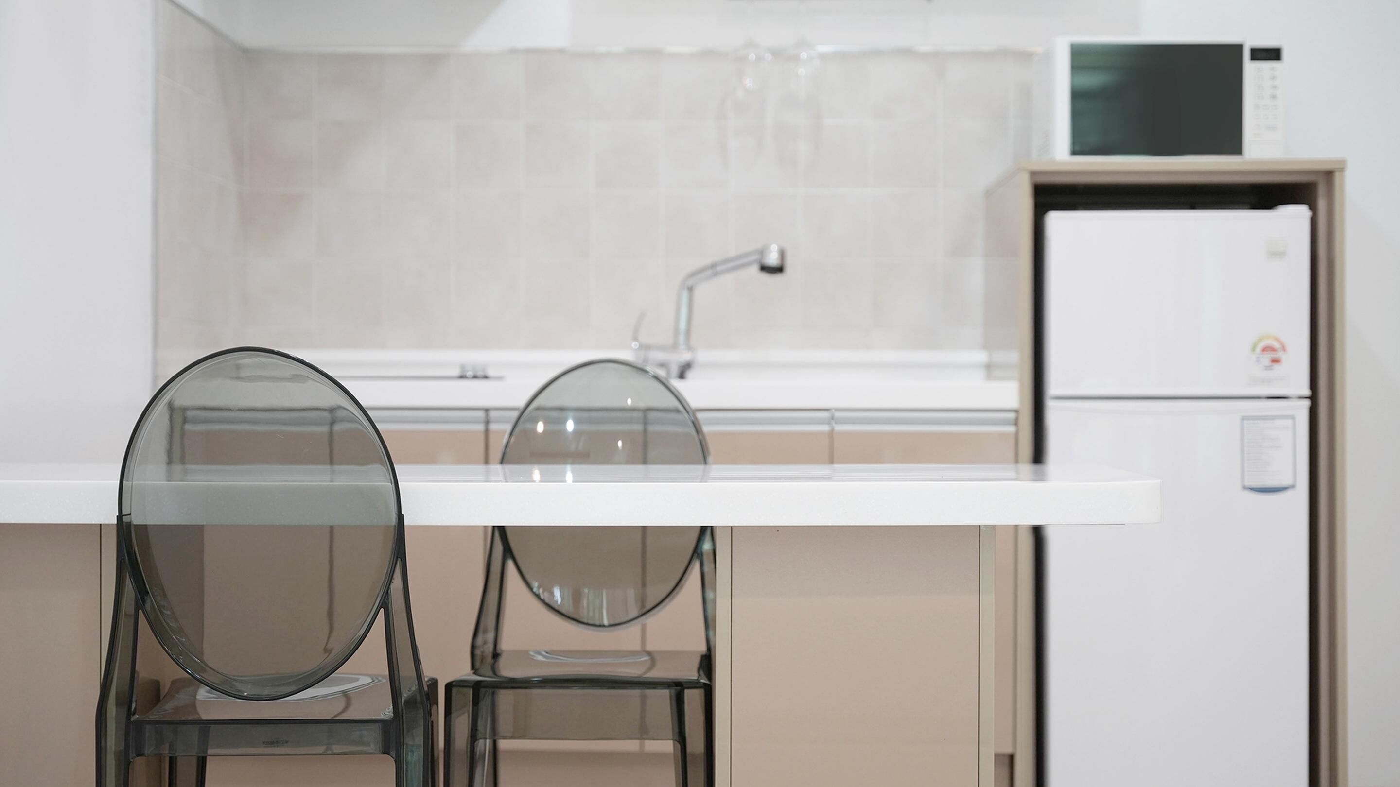 Una cocina moderna con encimera blanca, dos sillas de vidrio, un lavamanos de acero inoxidable y un refrigerador blanco.