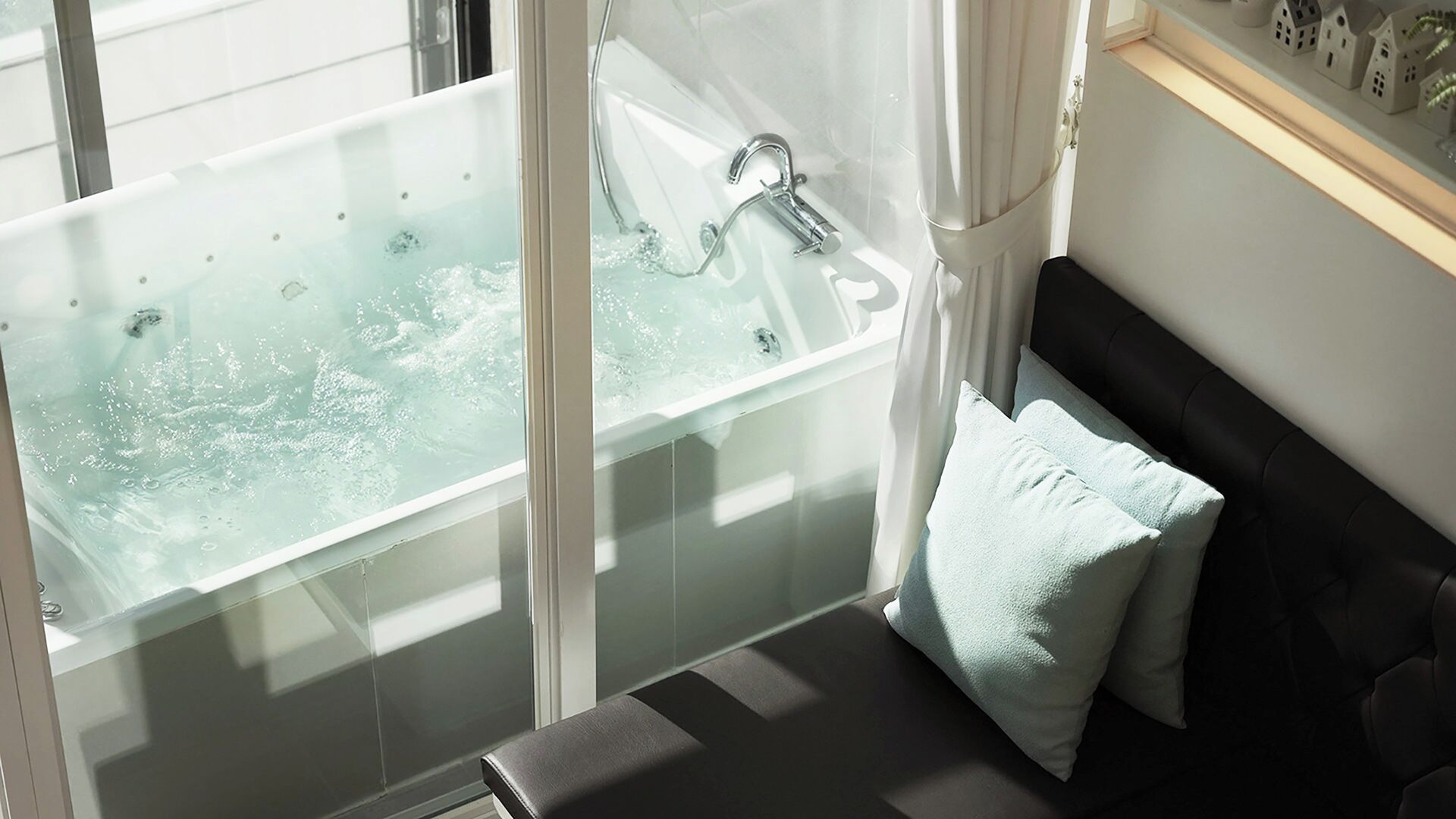A modern bathroom with a glass-enclosed bathtub, a black upholstered bench, and a white wall with a small shelf.