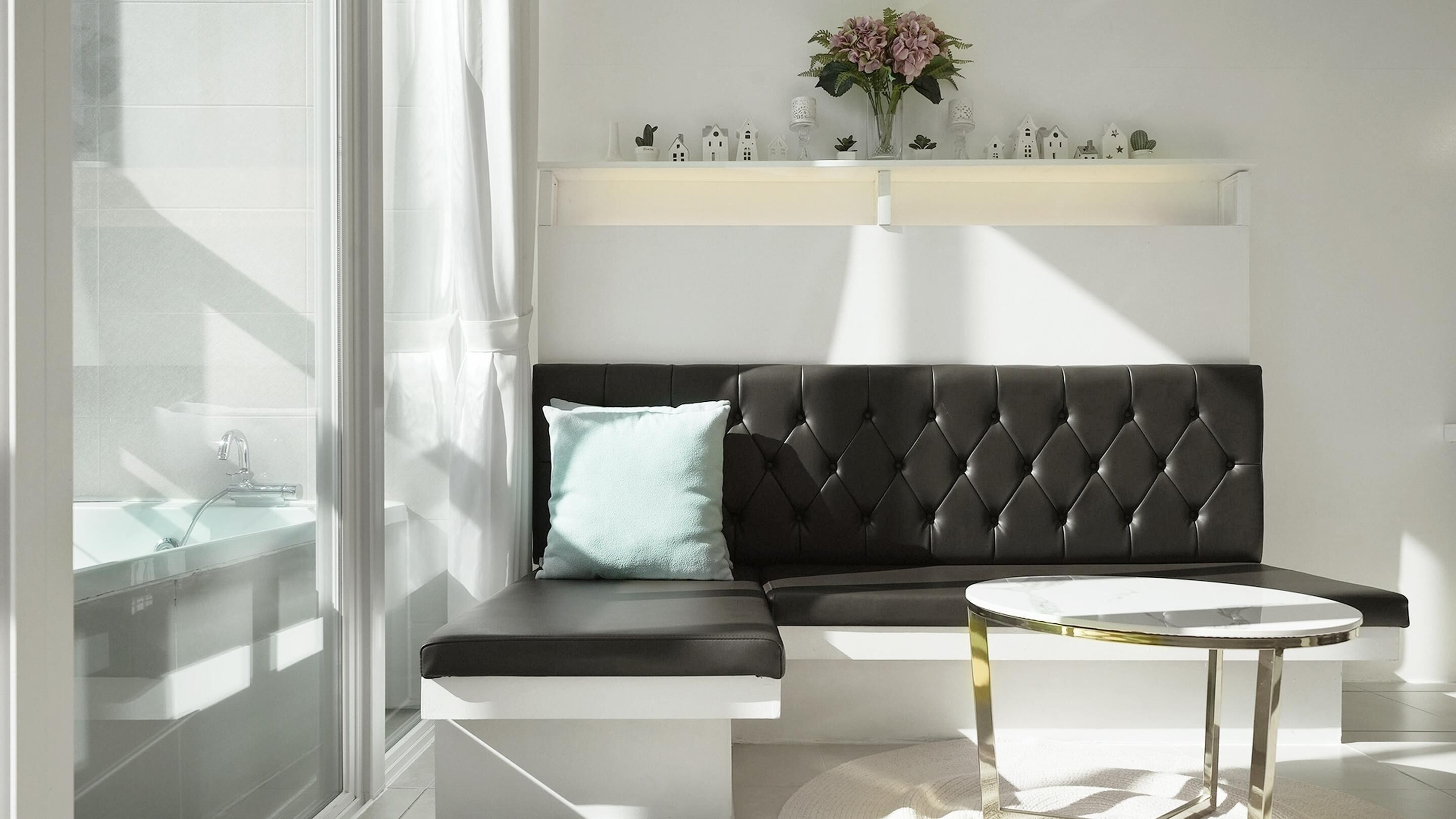 A modern living room with a black leather sofa, a round glass coffee table, and a white fireplace mantel with decorative items.