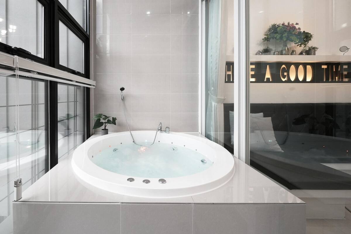 Une salle de bain moderne avec une grande baignoire, une douche en verre et une vue sur un lit où est posée une pancarte indiquant : « PASSEZ UN BON MOMENT ».