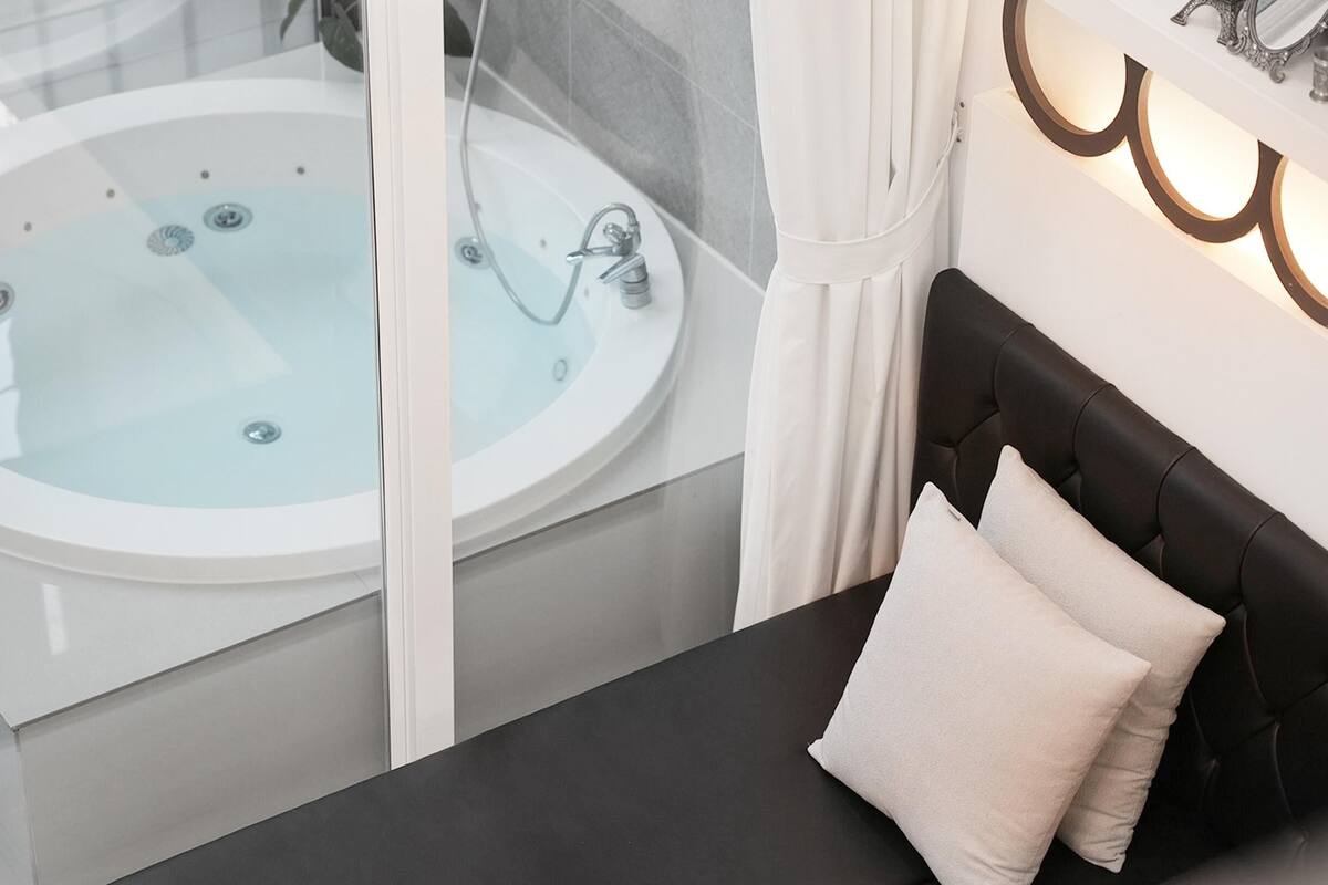 Une salle de bain moderne avec une baignoire sur pieds, une douche à parois de verre et une tête de lit noire recouverte de tissu, agrémentée d’un coussin blanc.