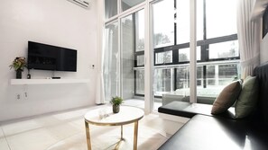 Un salon moderne doté d’un téléviseur à écran plat, d’une table basse ronde, d’un canapé avec des coussins et de grandes fenêtres offrant une vue sur l’extérieur.