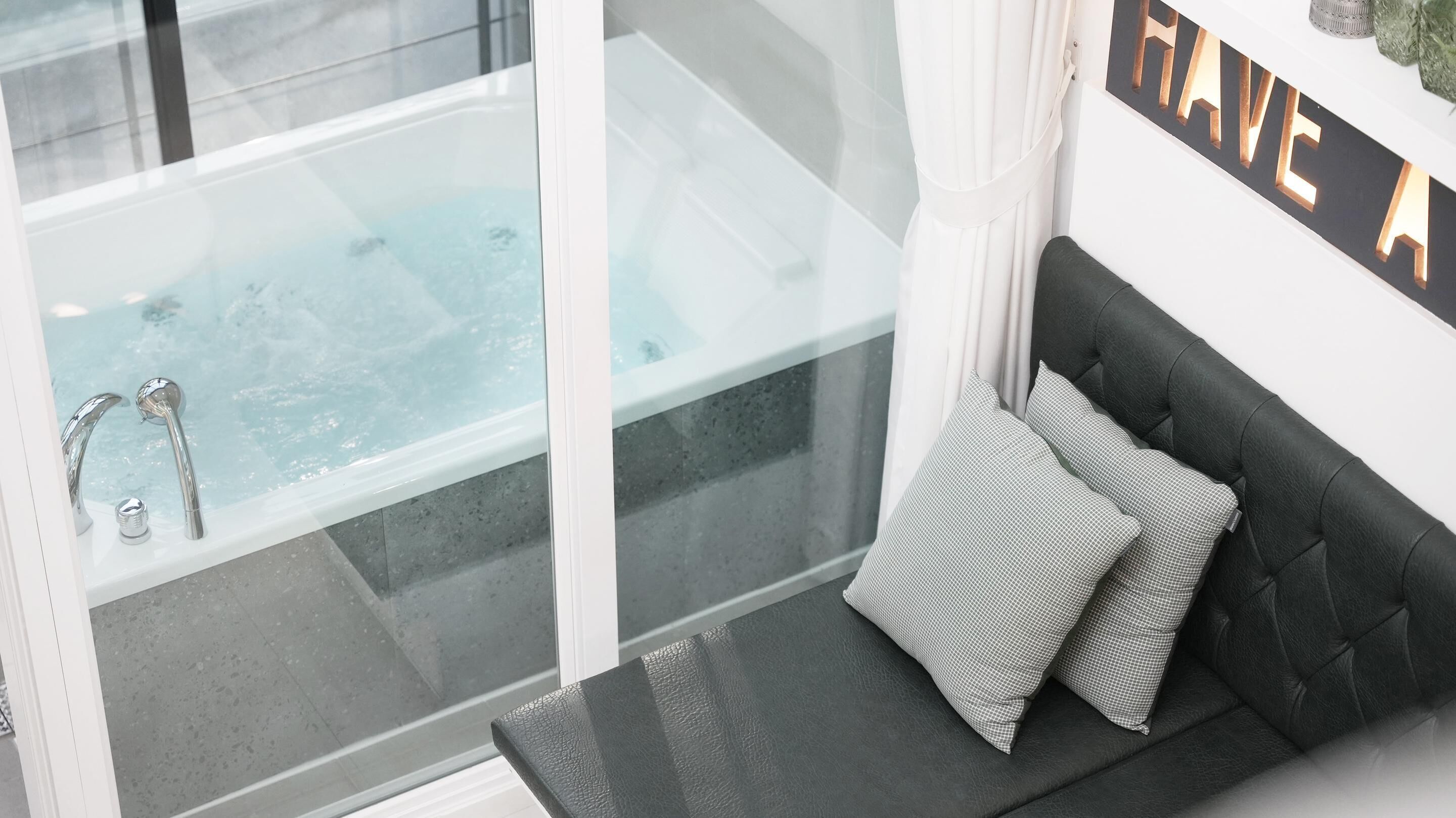 A modern bathroom with a glass-enclosed bathtub, a black leather sofa with pillows, and a 'HAVE A' sign on the wall.