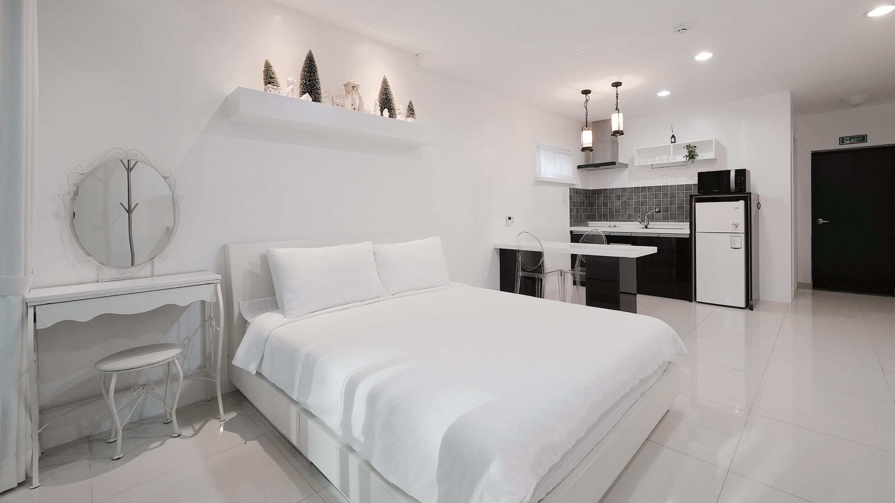 A modern, white-themed bedroom with a bed, a desk with a chair, a small table, and a mirror. There is a kitchenette with a sink and a refrigerator.