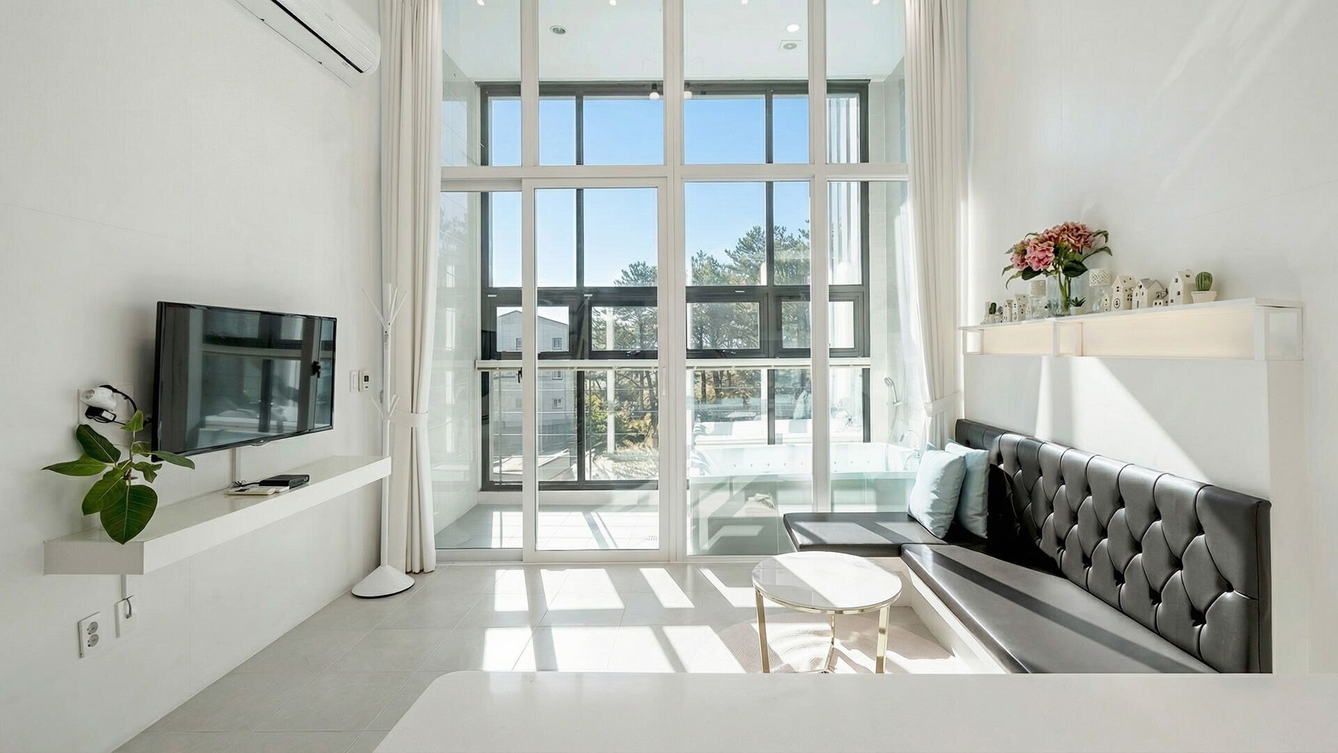Un salon moderne doté d’une grande porte vitrée donnant sur un balcon, d’un téléviseur à écran plat fixé au mur, d’un canapé gris et d’une petite table ronde.