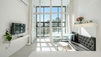 A modern living room with a large glass door leading to a balcony, a flat-screen TV mounted on the wall, a grey sofa, and a small round table.