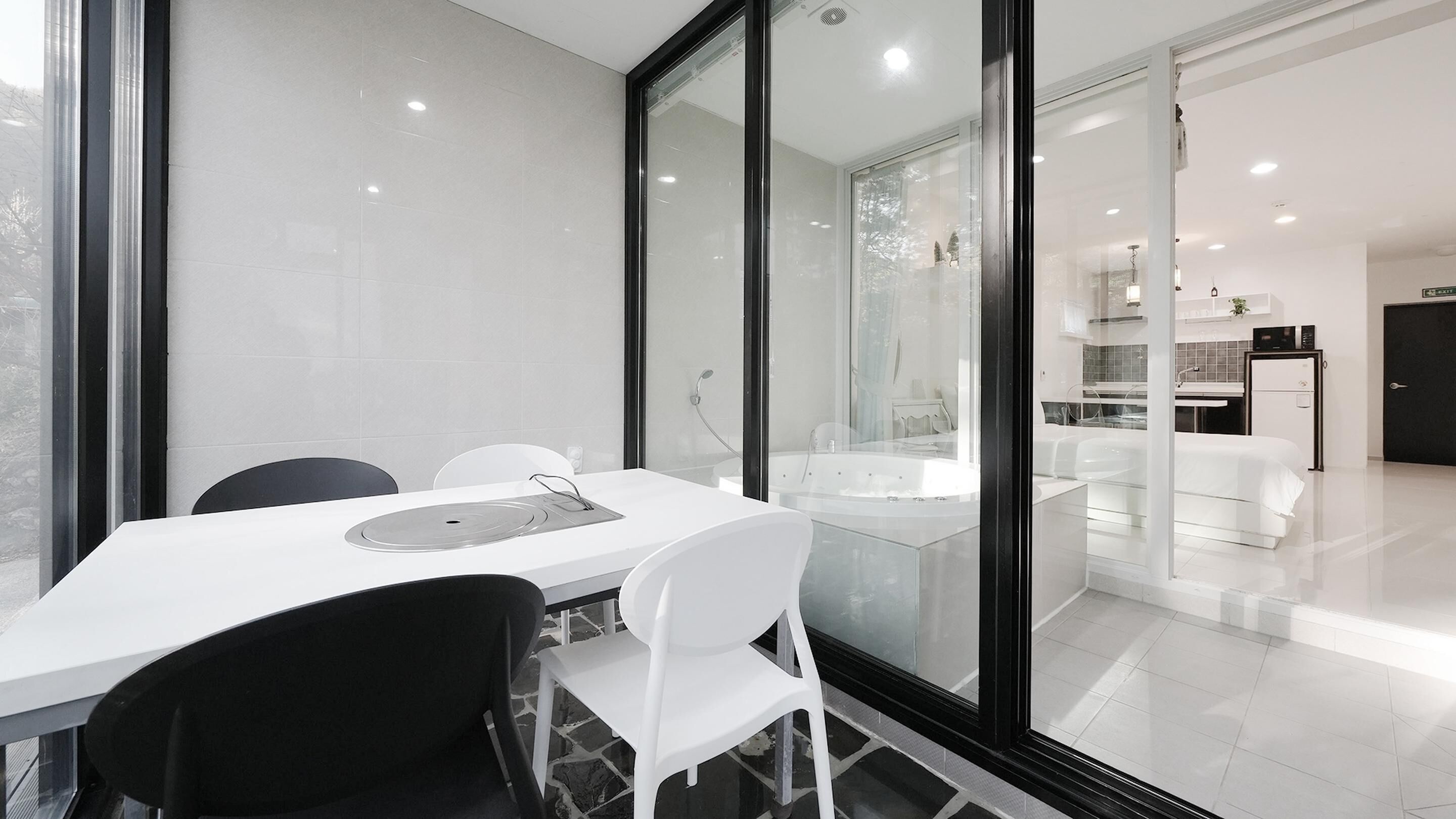 Une pièce moderne et bien éclairée, dotée d’un grand mur en verre, d’une table à manger blanche avec des chaises, et d’un espace cuisine visible au fond.
