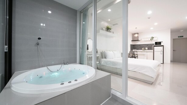 Ein modernes Badezimmer mit einer großzügigen Badewanne, einer Duschkabine aus Glas und einem sichtbaren Schlafzimmer mit Bett und einer kleinen Küchenzeile.