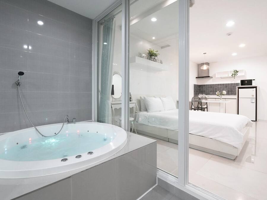 Ein modernes Badezimmer mit einer großzügigen Badewanne, einer Duschkabine aus Glas und einem sichtbaren Schlafzimmer mit Bett und einer kleinen Küchenzeile.