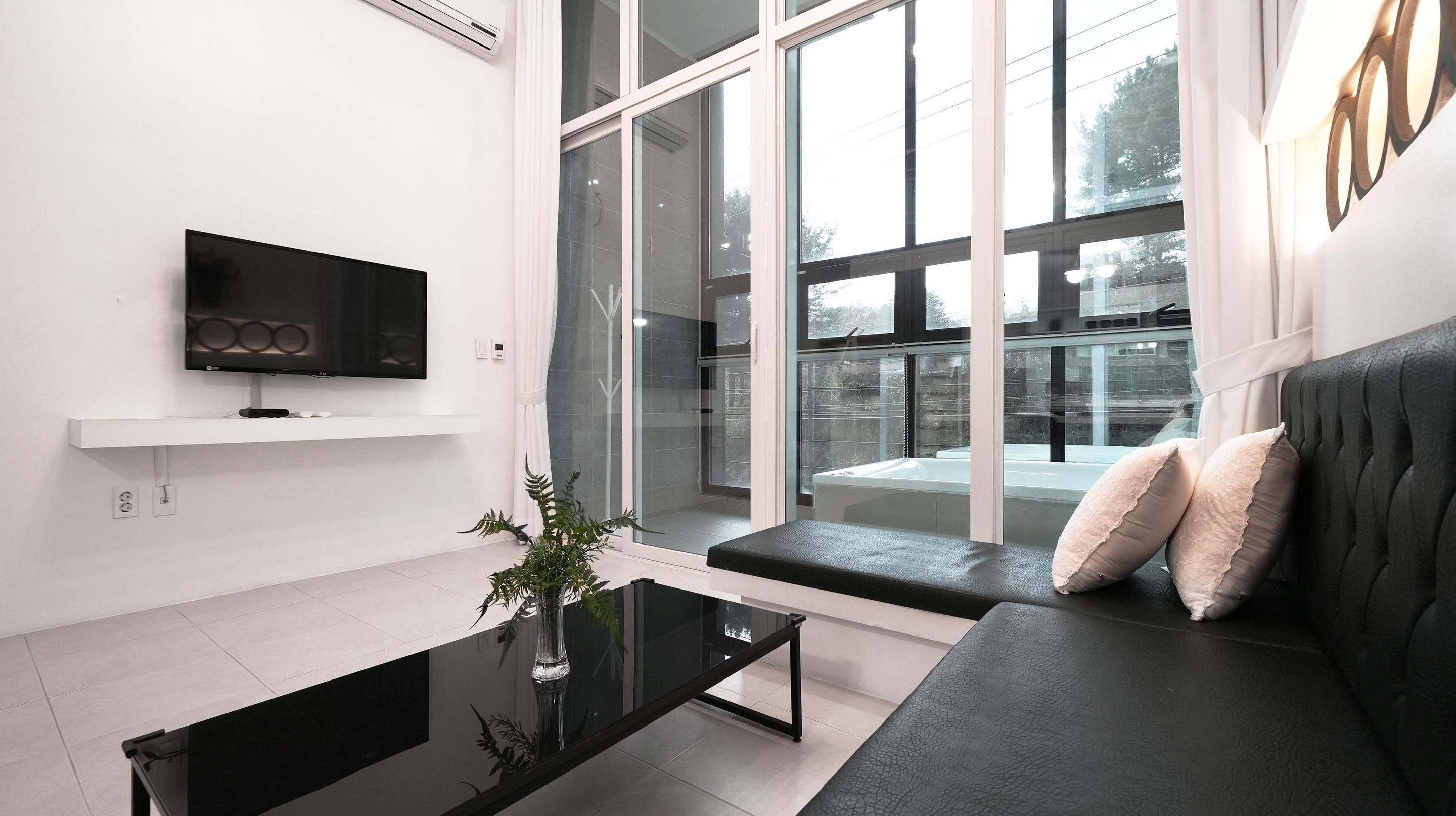 A modern living room with a black leather sofa, a glass coffee table, a flat-screen TV mounted on the wall, and a large window offering a view of the outdoors.