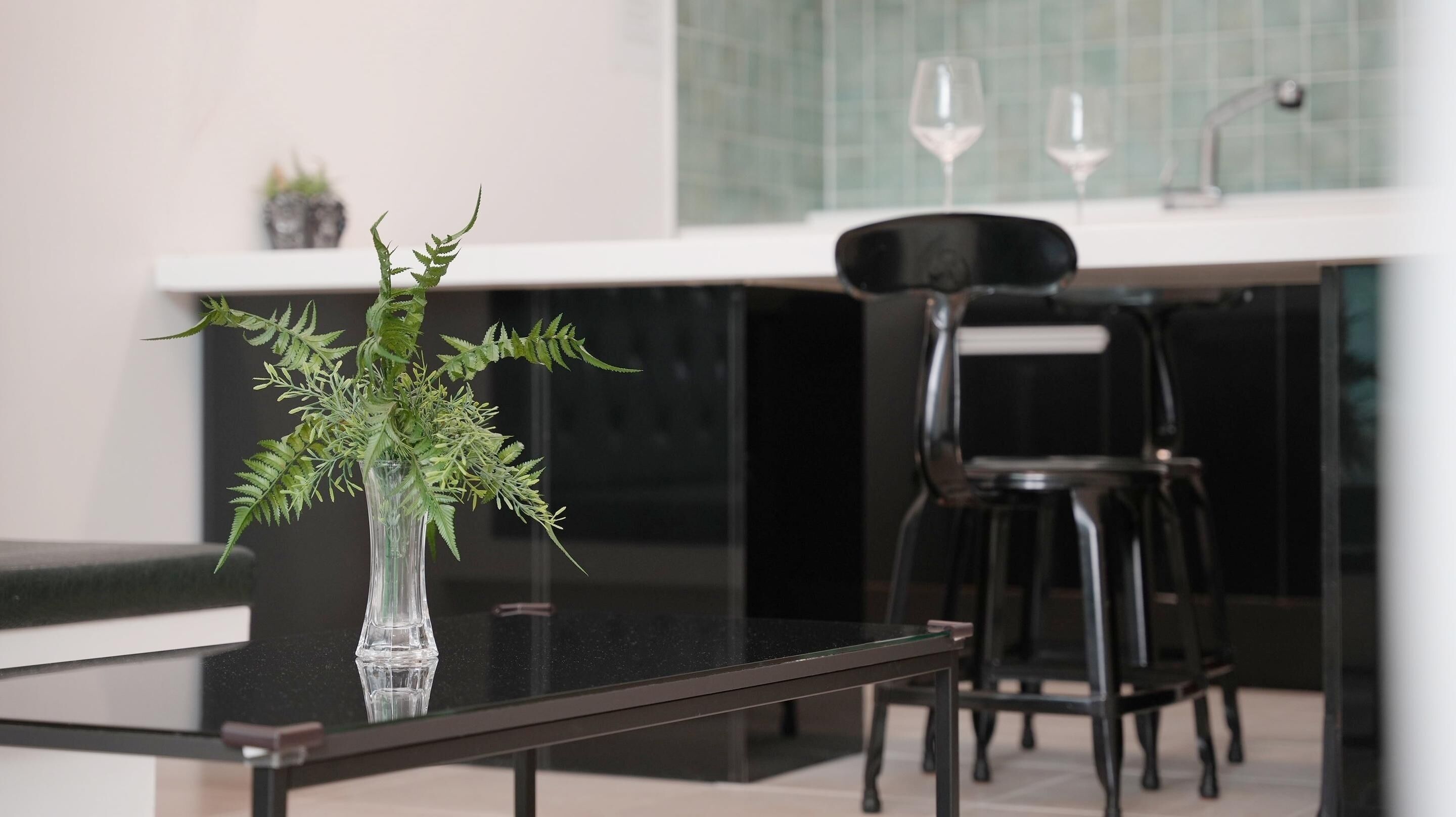 A modern kitchen with a glass table, a clear vase with green plants, and black chairs.