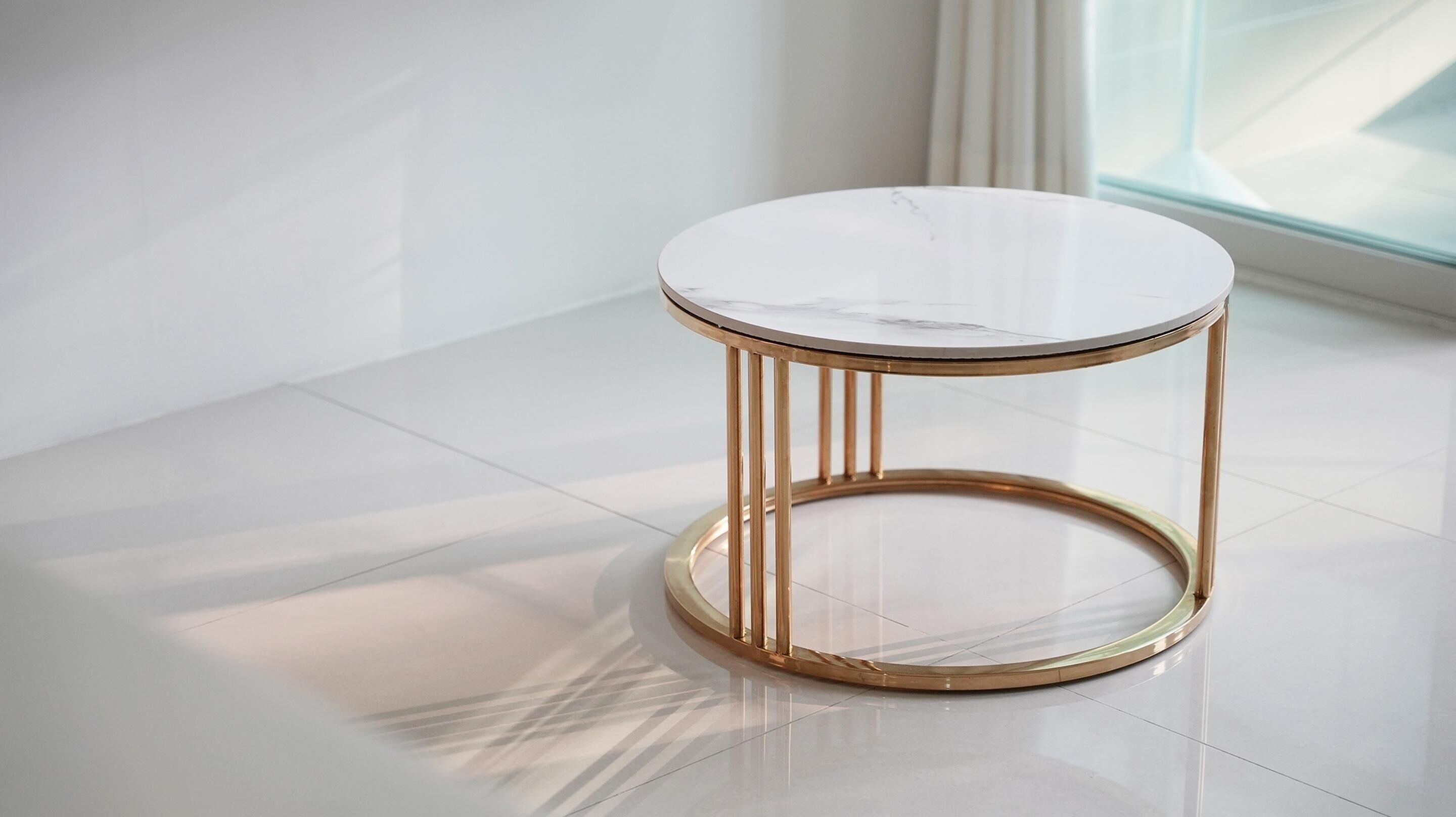 A round table with a marble top and gold legs, placed on a tiled floor in a bright room with a window.