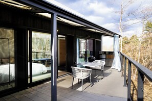 Una terraza moderna al aire libre con un dormitorio envuelto en vidrio, un comedor con sillas y vistas al bosque circundante.