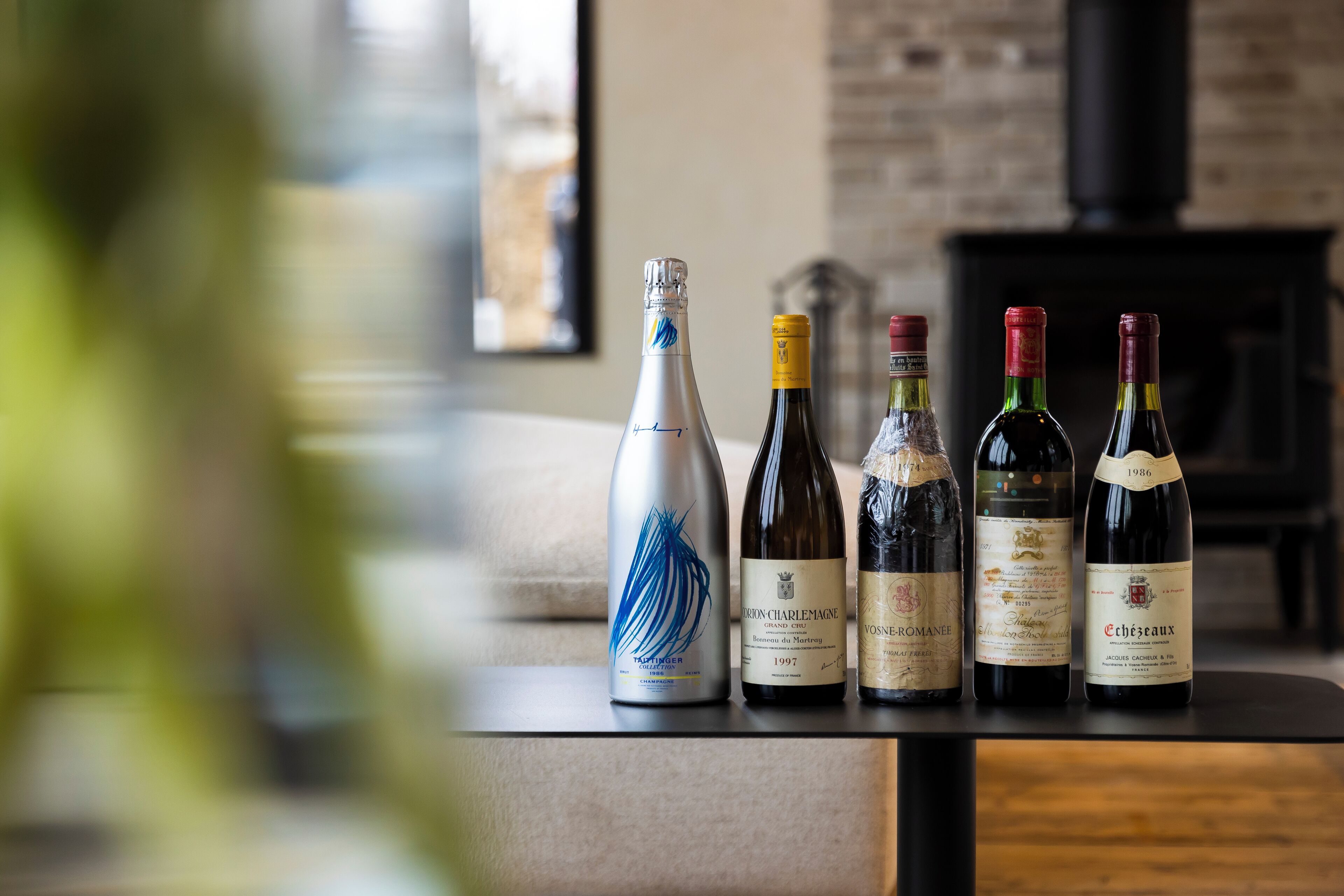 Quatre bouteilles de vin sont posées sur une table dans une pièce dont un mur est en briques et qui contient une cheminée.