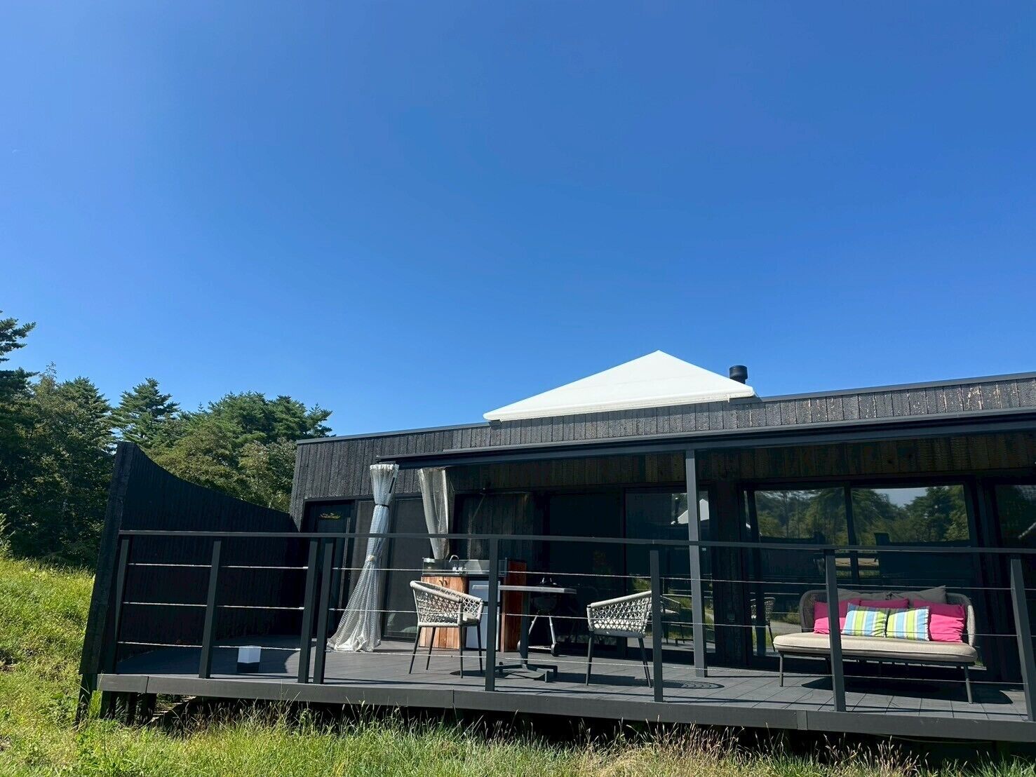 A modern building with a flat roof, large windows, and a covered outdoor seating area with two chairs and a sofa.
