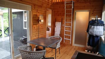 A wooden cabin interior with a dining table, wicker chairs, a ladder, a small kitchenette, and a sliding glass door leading to an outdoor deck.