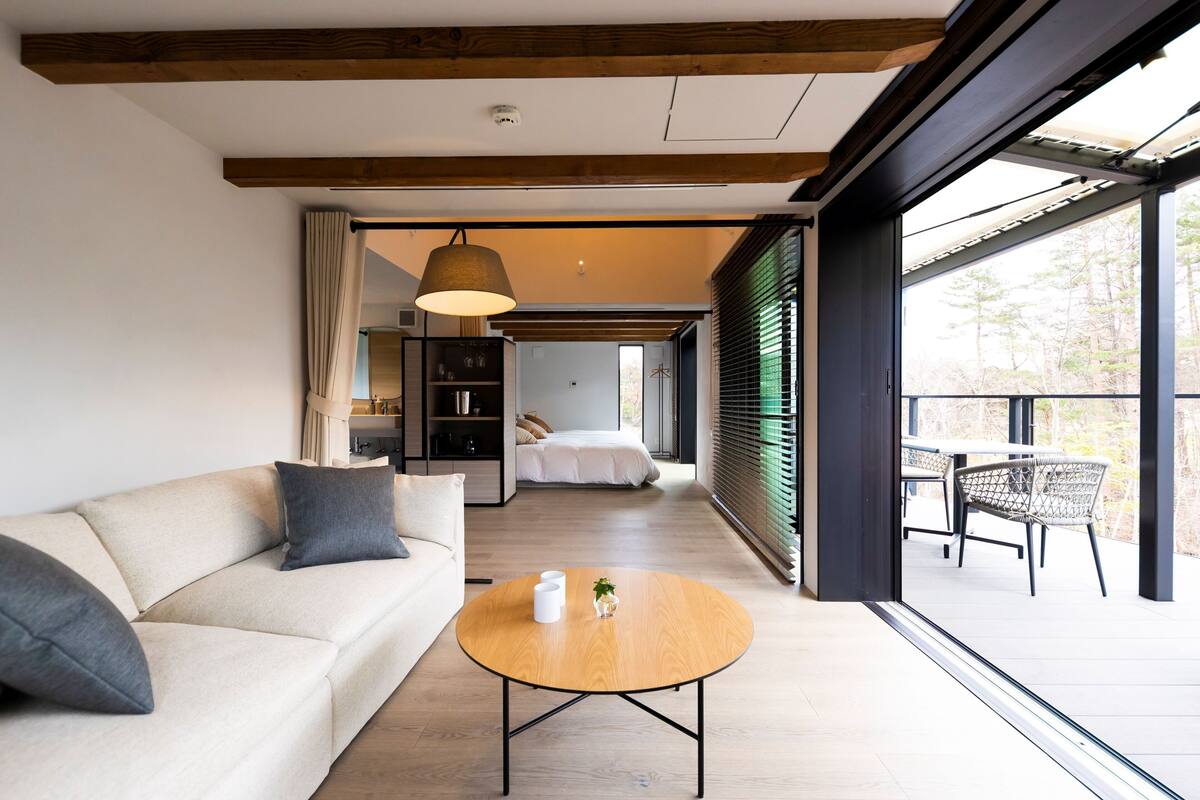 A modern interior with a sofa, round wooden table, and a view of a balcony with outdoor furniture.