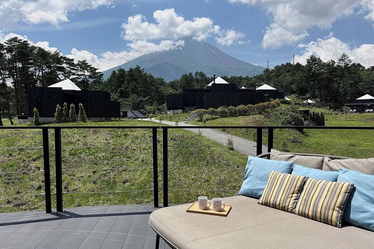 A deck with a sofa and a tray of mugs, overlooking a mountain and a forested area.