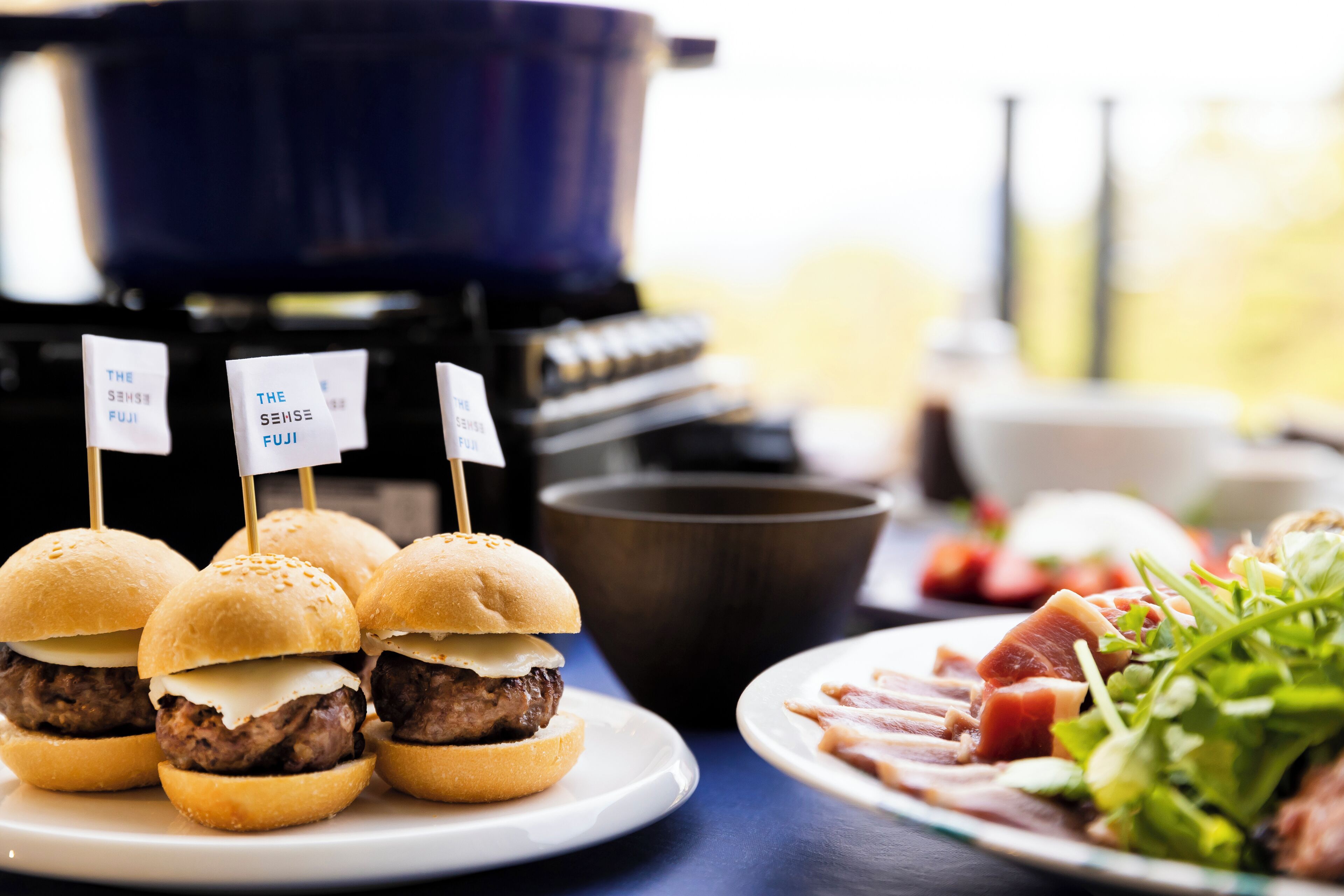A table with three mini burgers on a plate, a bowl of salad, and a grill in the background.