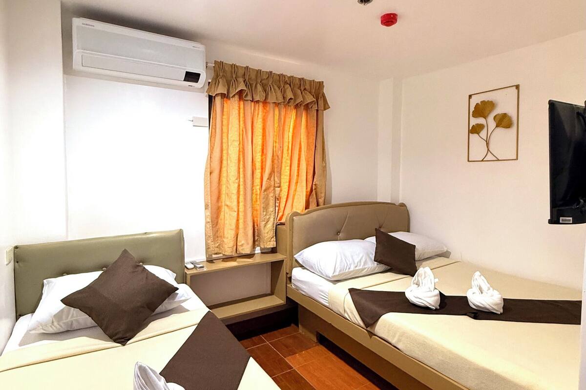 A hotel room with two beds, a nightstand, an air conditioning unit, a window with curtains, and a wall-mounted TV.