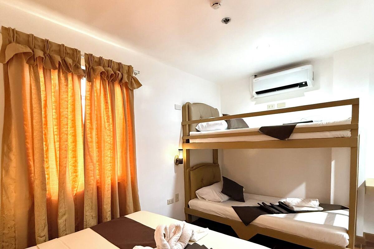 A hotel room with a bunk bed, a single bed, a window with curtains, an air conditioning unit, and a tiled floor.