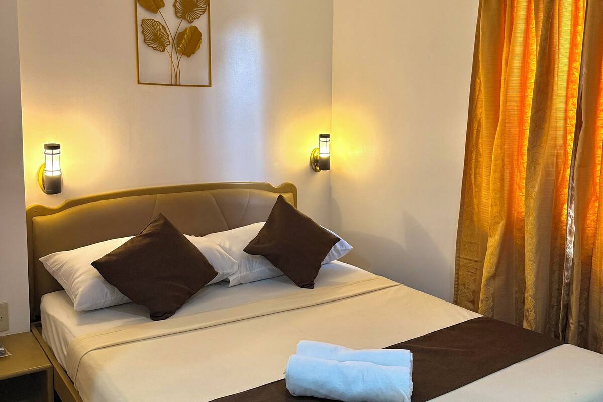 A neatly made bed with a brown headboard, two bedside lamps, and a wall-mounted artwork of leaves.