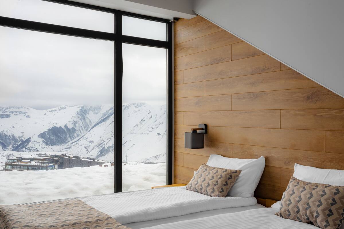 Un dormitorio con un gran ventanal, cabecera de madera y cama con almohadas estampadas.
